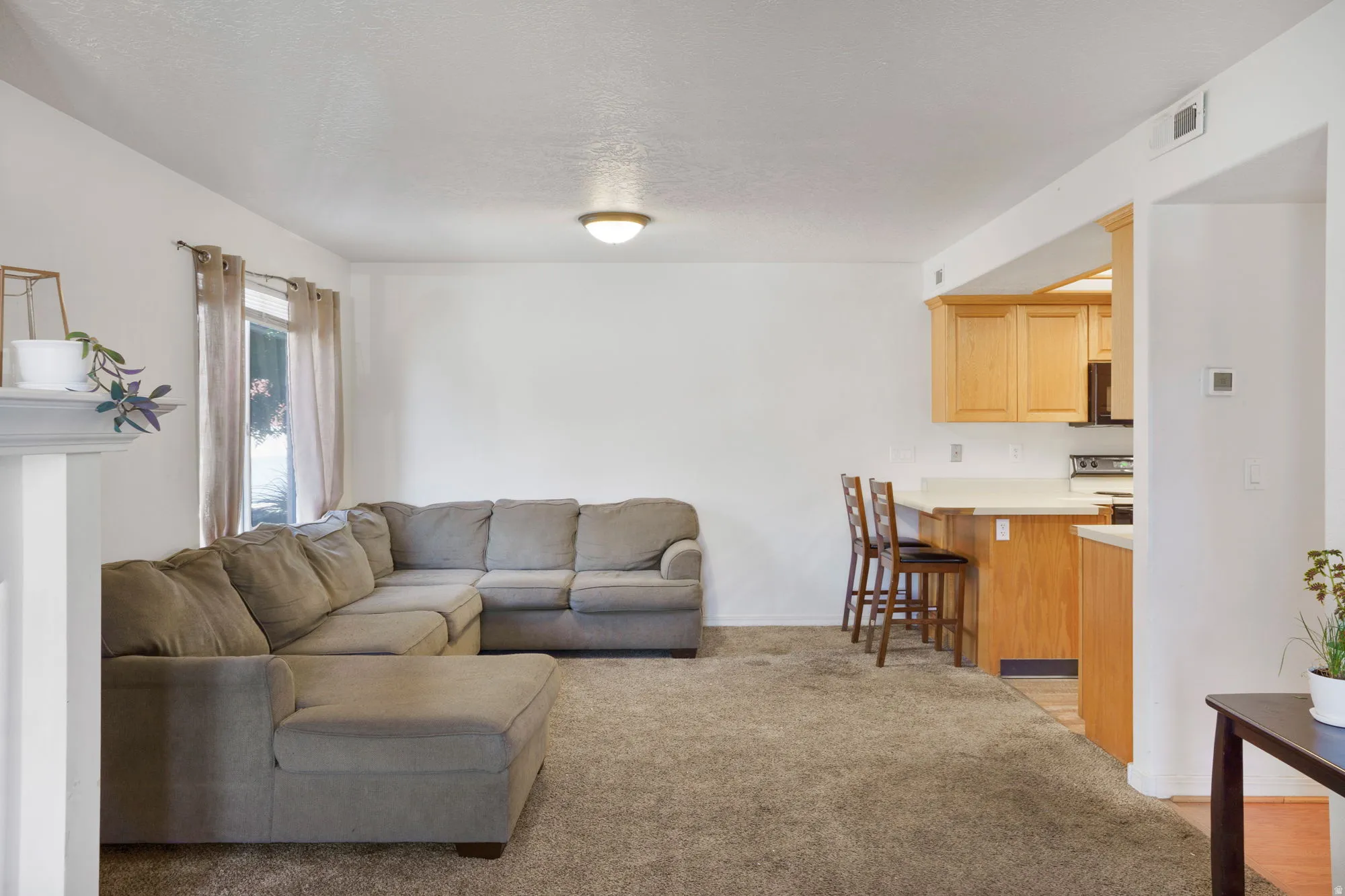 Open-concept living area featuring a carpeted floor, a window with curtains, a built-in light fixture, and a partial view of a kitchen bar area with light wood cabinetry and a white countertop