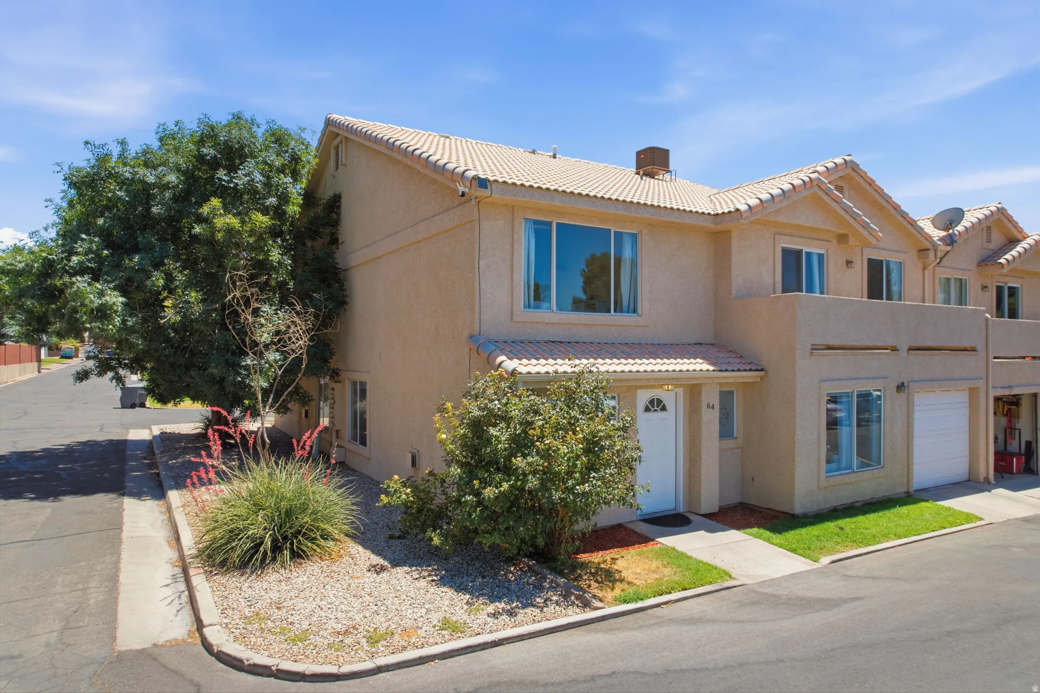 Tan stucco exterior with a tiled roof, featuring a private front entrance, an integrated garage, a second-story balcony, and mature landscaping