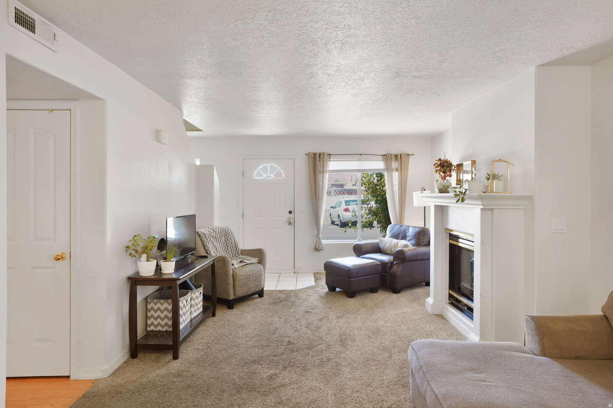 Carpeted living area featuring a white fireplace with a mantel, a front entry door with a decorative glass insert, a window with curtains, and white paneled doors