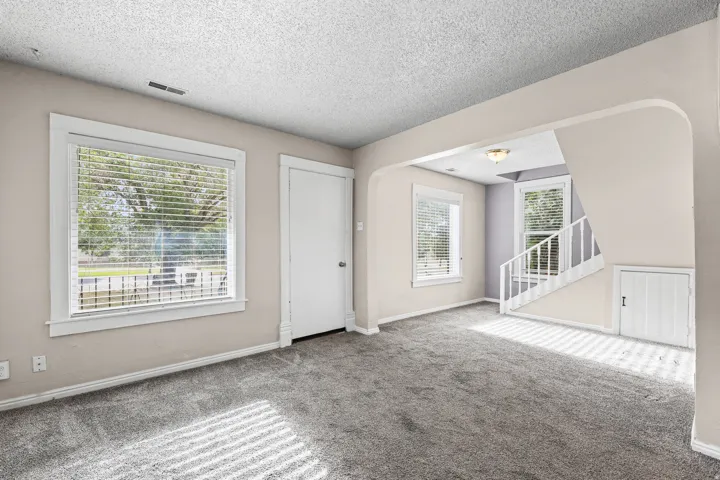 Inviting interior featuring new gray carpet, neutral wall tones, multiple windows with blinds, a prominent arched doorway, and white staircase with matching balusters