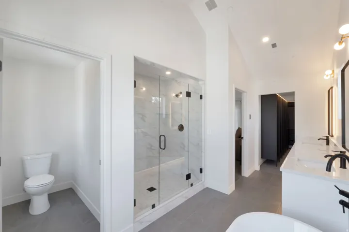 Spacious bathroom featuring a glass-enclosed shower with tile surround, dual vanity with undermount sinks and dark fixtures, and light gray floor tiles