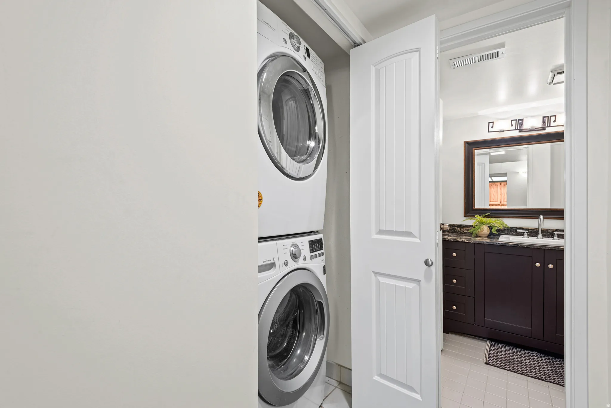 Stackable laundry appliances within a dedicated closet, with a paneled interior door opening to a bathroom featuring a dark wood-finish vanity, a stone countertop, an undermount sink, and an ornamental framed mirror