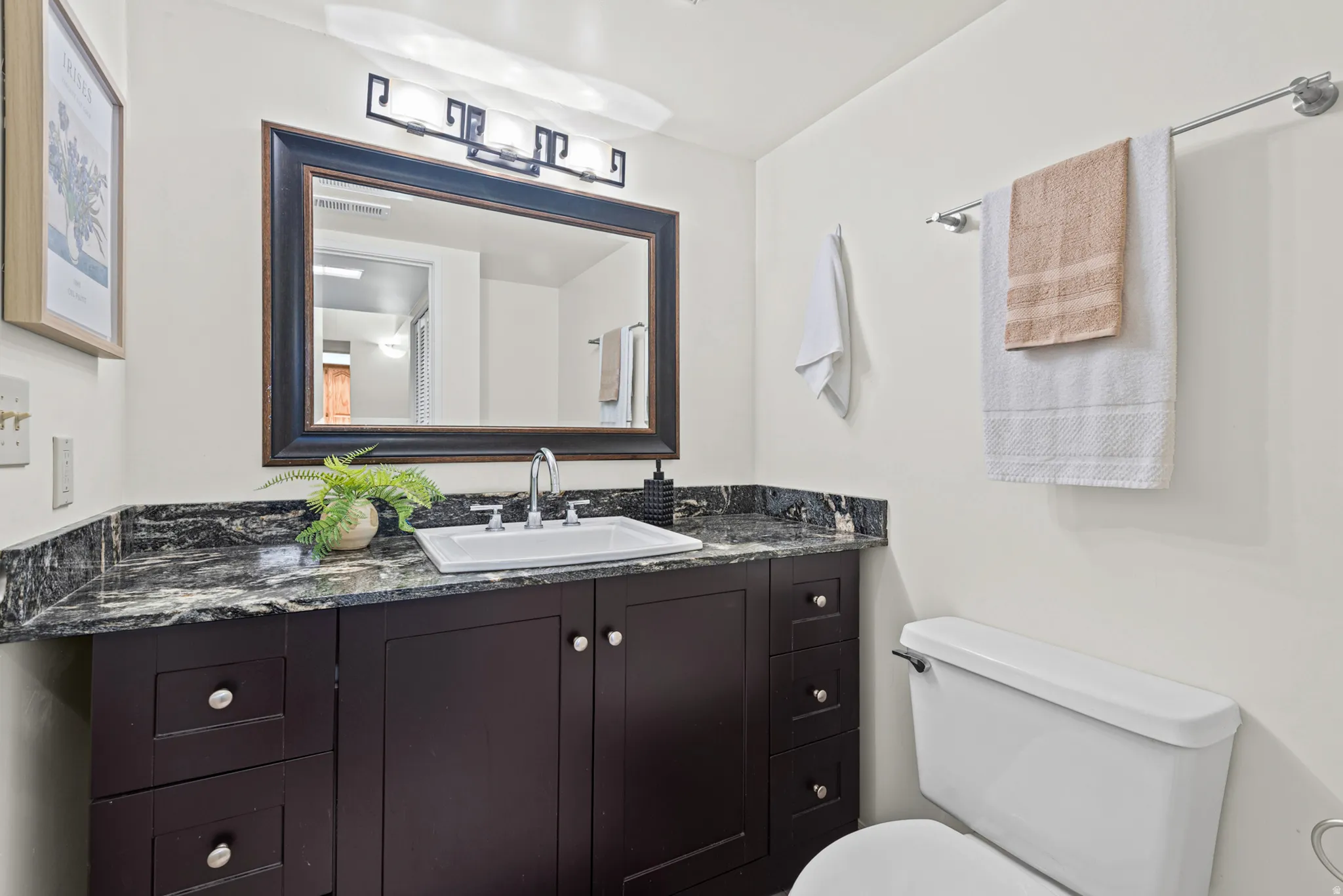 Dark wood-finish vanity with a stone countertop, an integrated rectangular sink, a framed mirror, and a three-light vanity fixture