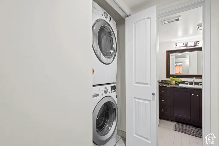Stackable laundry appliances within a dedicated closet, with a paneled interior door opening to a bathroom featuring a dark wood-finish vanity, a stone countertop, an undermount sink, and an ornamental framed mirror