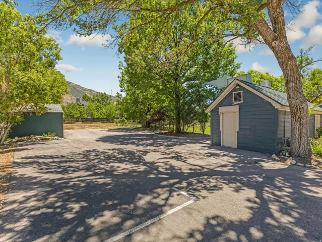 Paved parking area framed by mature trees, featuring a blue-sided outbuilding with a light-colored door and a dark green shed
