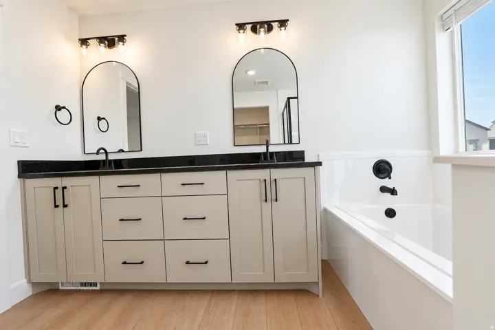 Dual vanity with black countertops, matte black fixtures, arched mirrors, and wood-finish flooring, complemented by a built-in bathtub