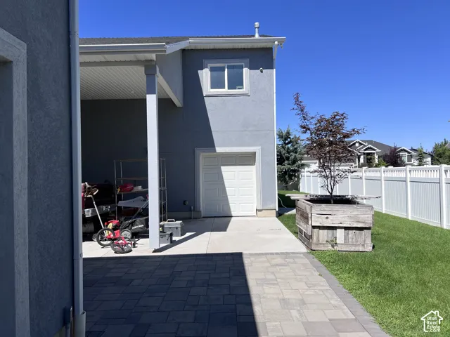 Paver patio with a covered outdoor area, detached garage, white vinyl fencing, and a mature tree in a wooden planter