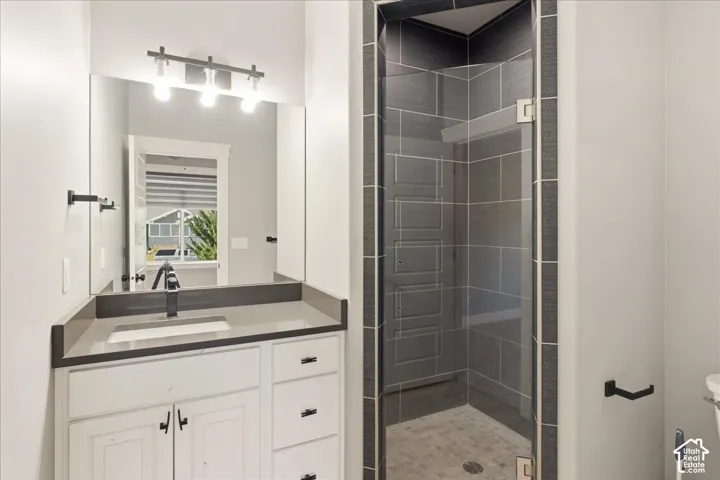 Bathroom featuring a glass-enclosed shower with dark tile surround, single vanity with light-toned countertop, white cabinetry with dark hardware, and a framed mirror with a horizontal light fixture
