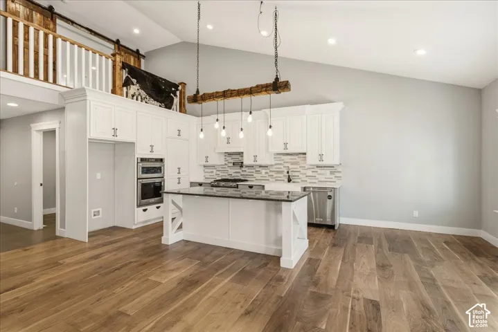 Open-concept kitchen featuring white cabinetry, a central island with a dark countertop, stainless steel appliances, and a unique pendant light fixture