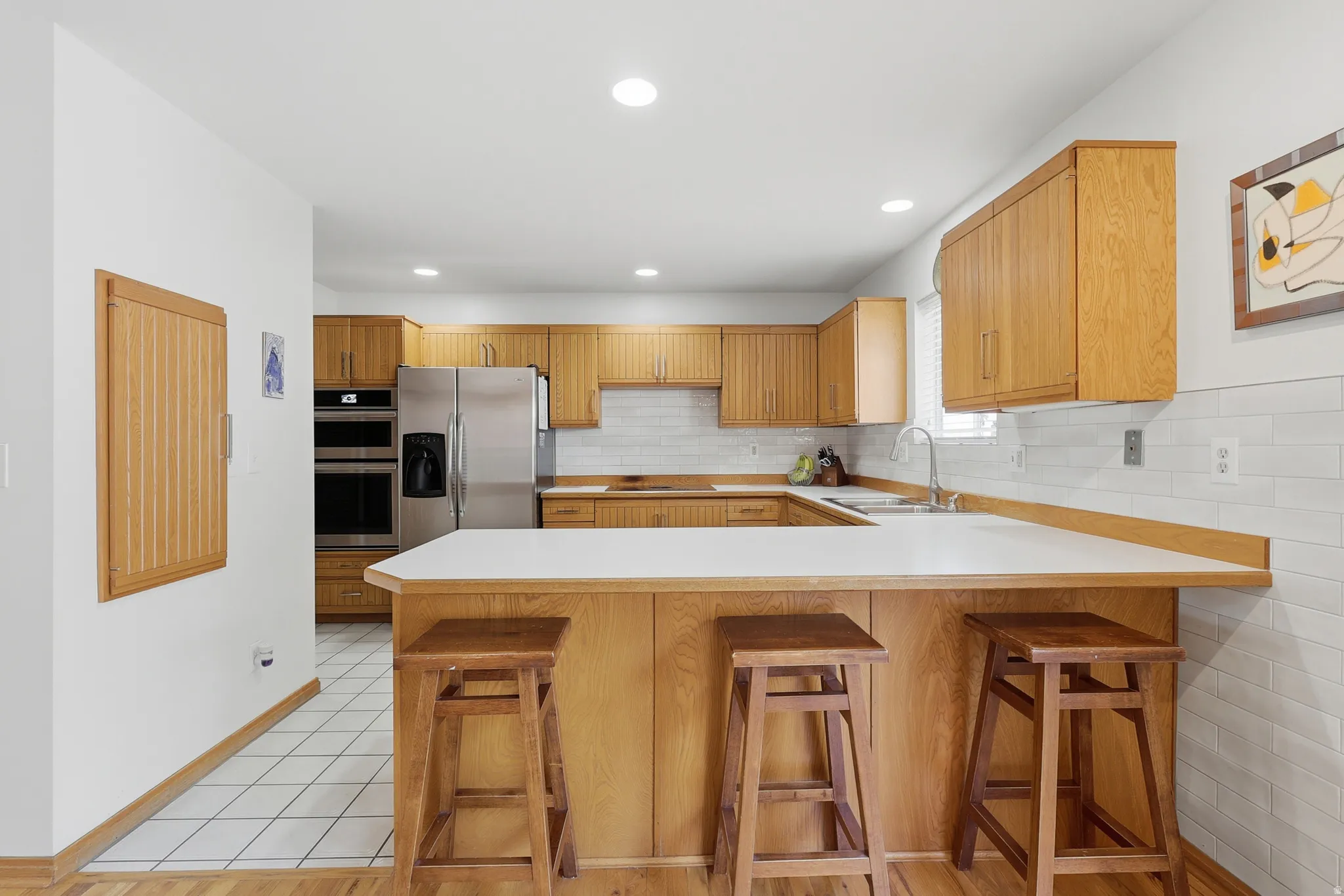 Kitchen featuring light wood cabinetry, white subway tile backsplash, stainless steel appliances, a double basin sink, and a peninsula with a white countertop