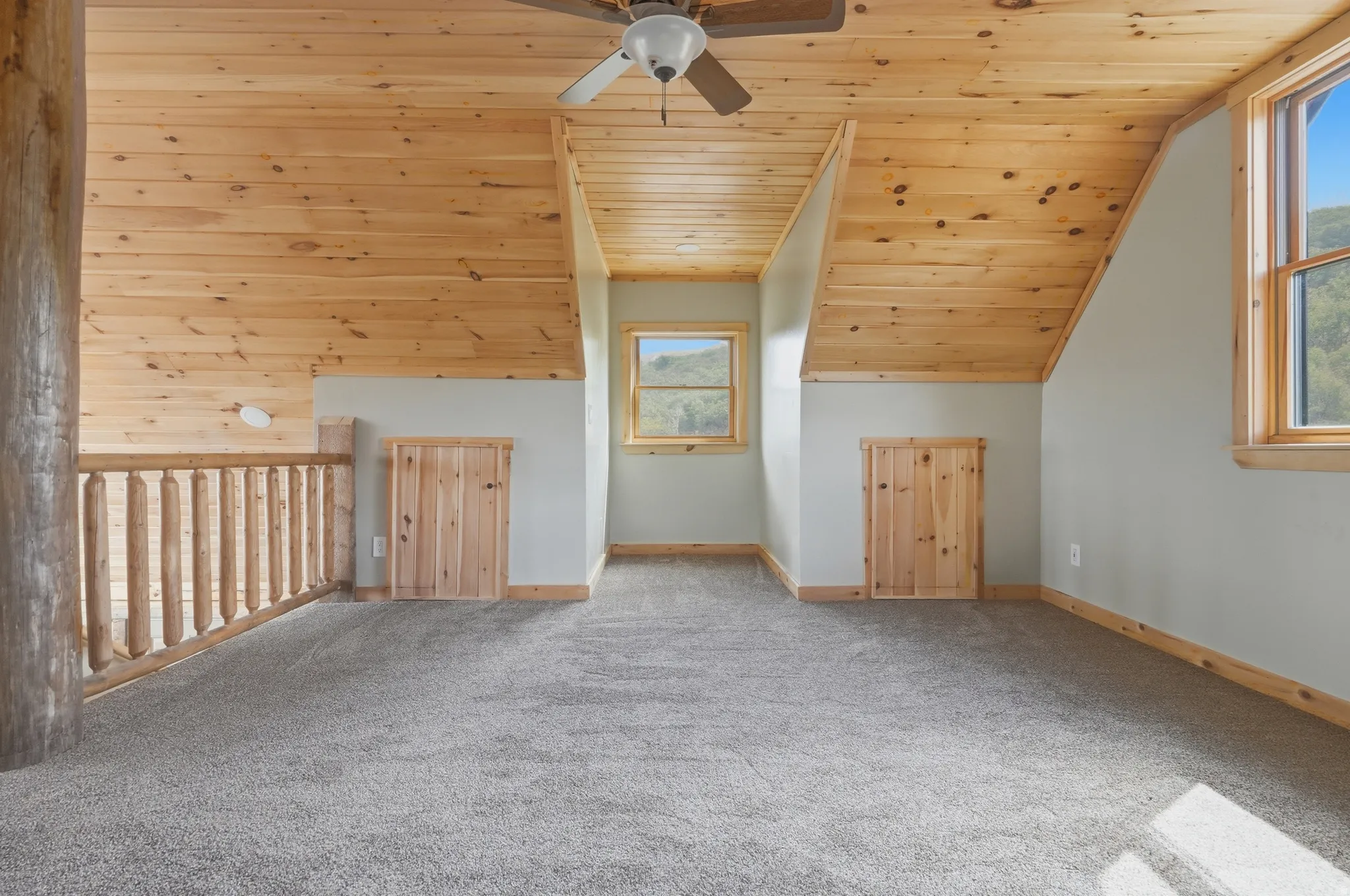 Loft space featuring sloped wood-finish ceilings, wood-finish window frames, two windows, two wood-finish storage access doors, and soft gray carpeting