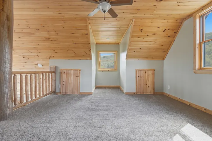 Loft space featuring sloped wood-finish ceilings, wood-finish window frames, two windows, two wood-finish storage access doors, and soft gray carpeting