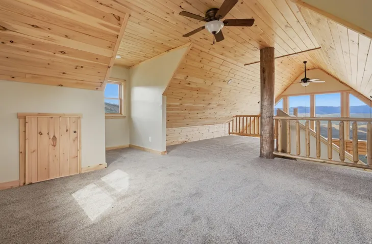 Spacious loft area featuring natural wood plank ceilings and trim, light-colored carpeting, multiple ceiling fans, and a large window wall offering panoramic outdoor scenery
