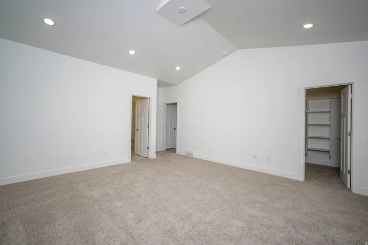 Spacious room featuring vaulted ceilings, recessed lighting, neutral carpeting, and multiple doorways including a walk-in closet with built-in shelving