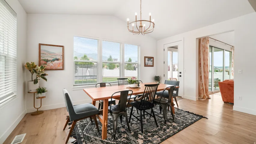 Bright dining area featuring wood-finish flooring, a contemporary chandelier, multiple windows with blinds, a white door, and an open doorway to another room with a sliding glass door