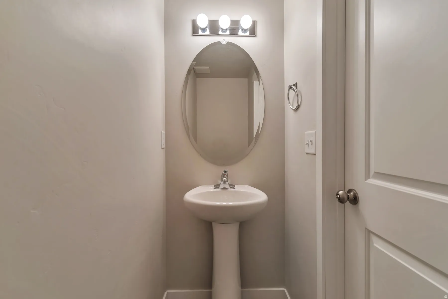 Pedestal sink with a chrome faucet, oval wall mirror, and a three-light vanity fixture