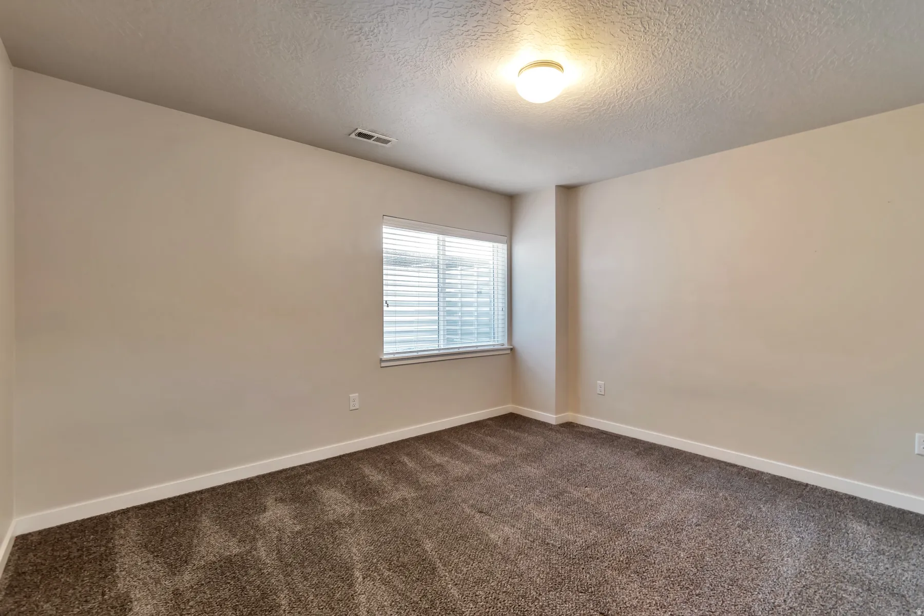 Neutral tone wall paint with white baseboards, textured ceiling with a flush-mount light fixture, and a window featuring horizontal blinds