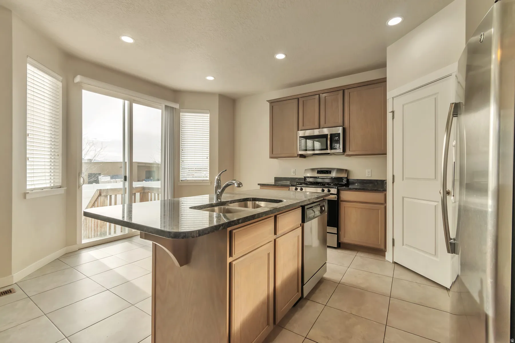 Kitchen featuring a central island with a double basin sink, stainless steel appliances, cabinetry with a natural wood finish, granite countertops, and tile flooring