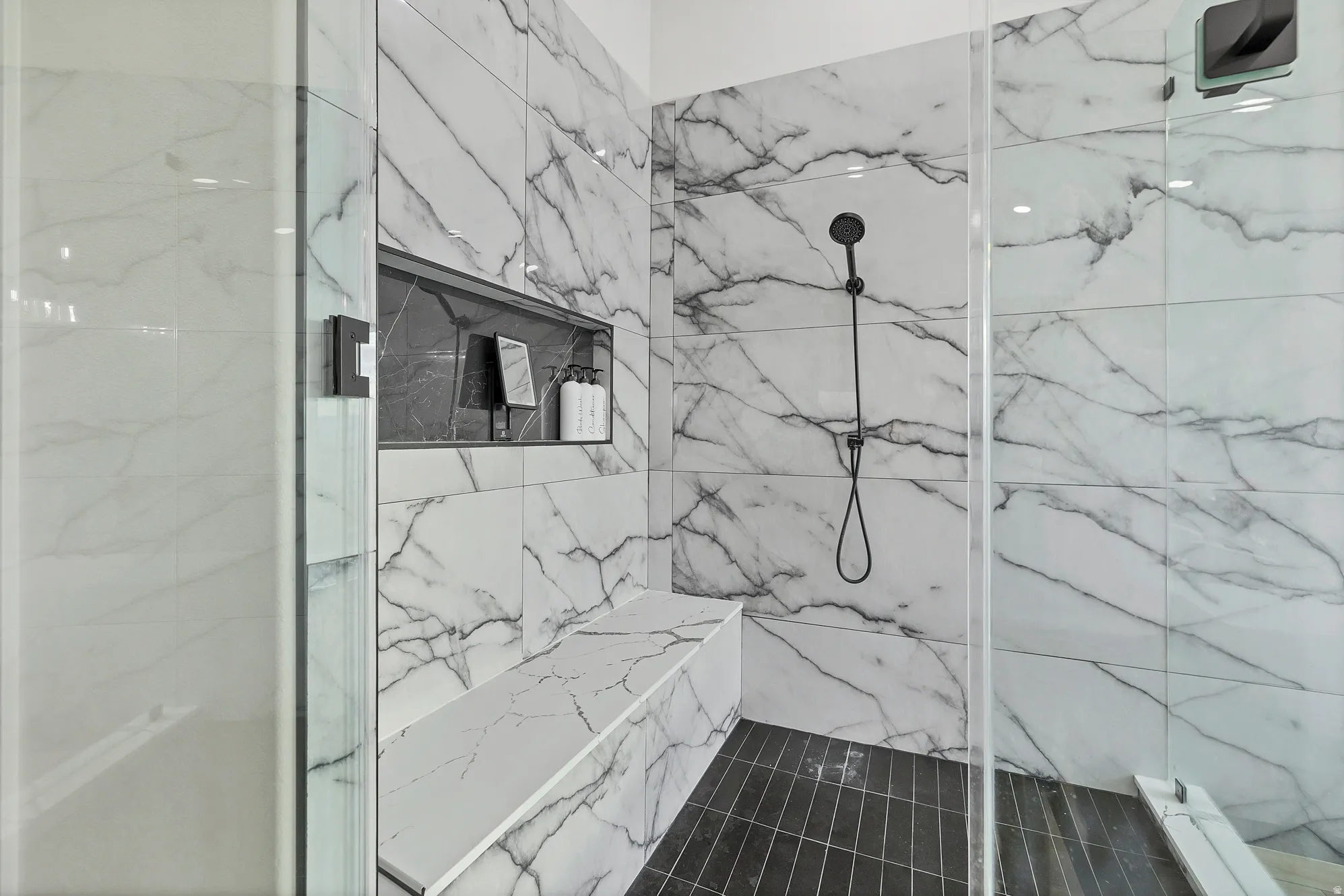 Walk-in shower featuring large format marble-look tile walls, a built-in shower bench, a recessed niche, dark tile flooring, and a matte black shower fixture