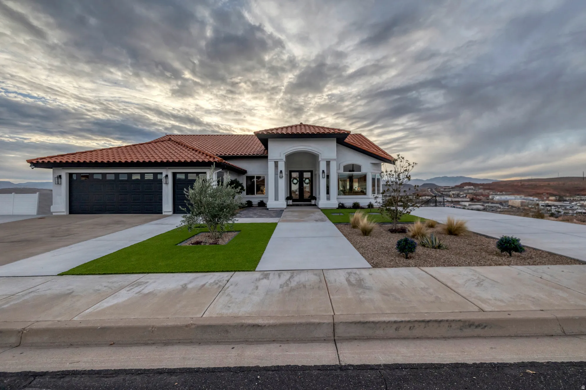 White stucco exterior with a terracotta tile roof, prominent arched entryway, and dual black garage doors