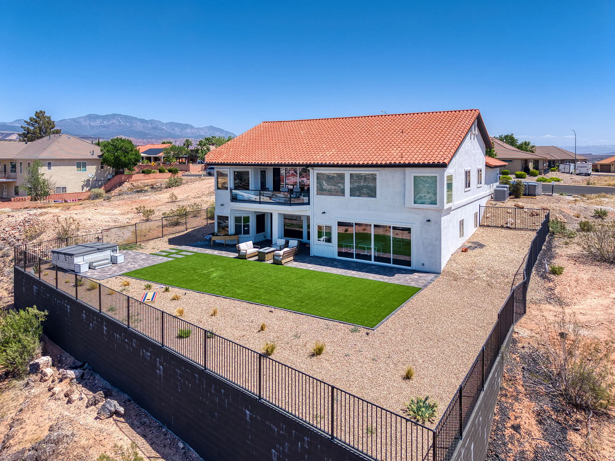Stucco exterior with a terra cotta tile roof, featuring a second-story balcony with glass railing, ground-level patio access, and a manicured synthetic lawn