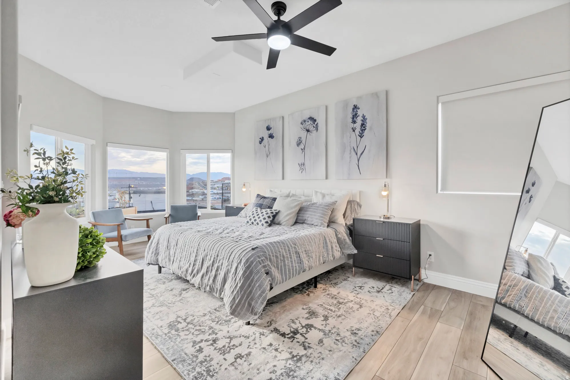 Spacious bedroom featuring light wood-finish flooring, a modern ceiling fan, and multiple windows with roller shades