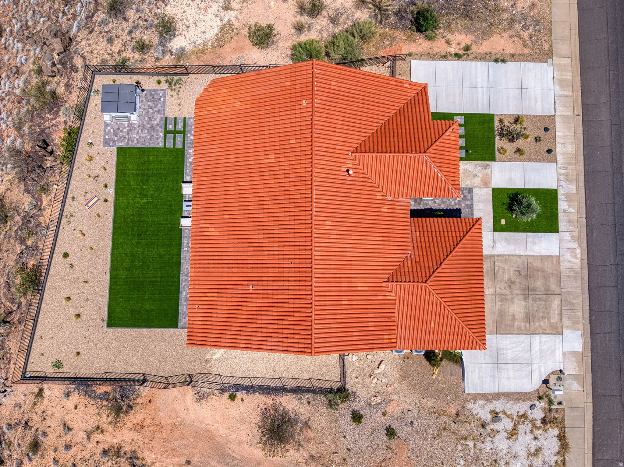 Aerial perspective showcasing a tiled roof, extensive concrete paving, a fenced perimeter, artificial turf, and a hot tub