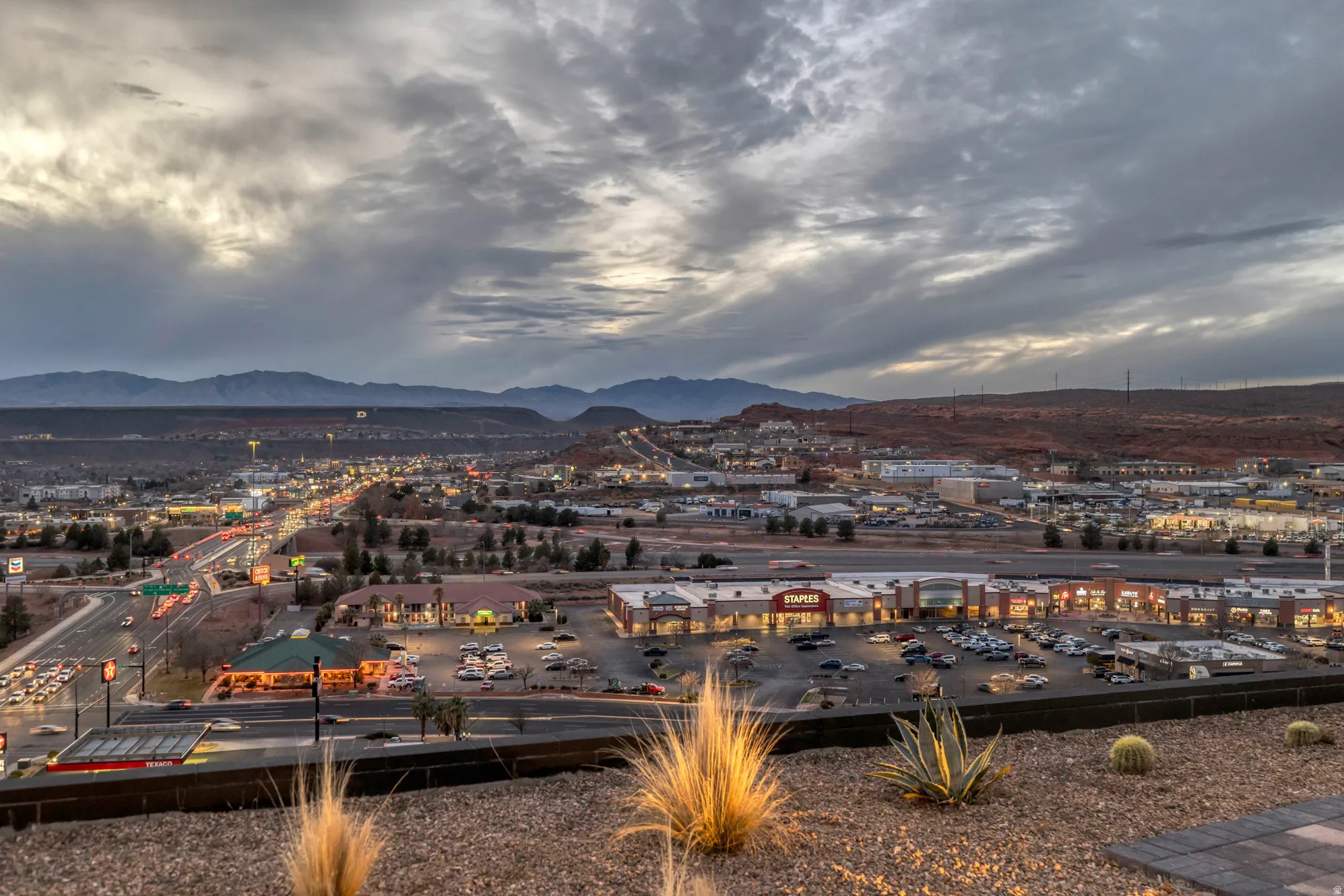 Elevated desert landscaping featuring agave and cacti, overlooking a commercial center with ample parking, and an expansive cityscape with distant mountains