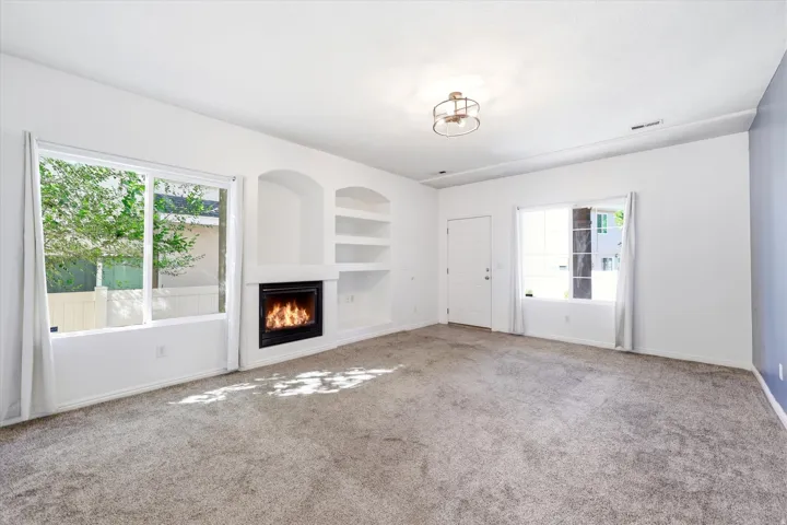 Integrated fireplace with built-in shelving, two large windows, white walls, and gray carpet flooring