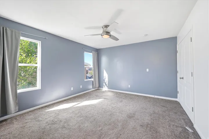 Spacious room featuring gray wall paint, two windows with gray drapes, gray carpet flooring, and a ceiling fan with integrated lighting