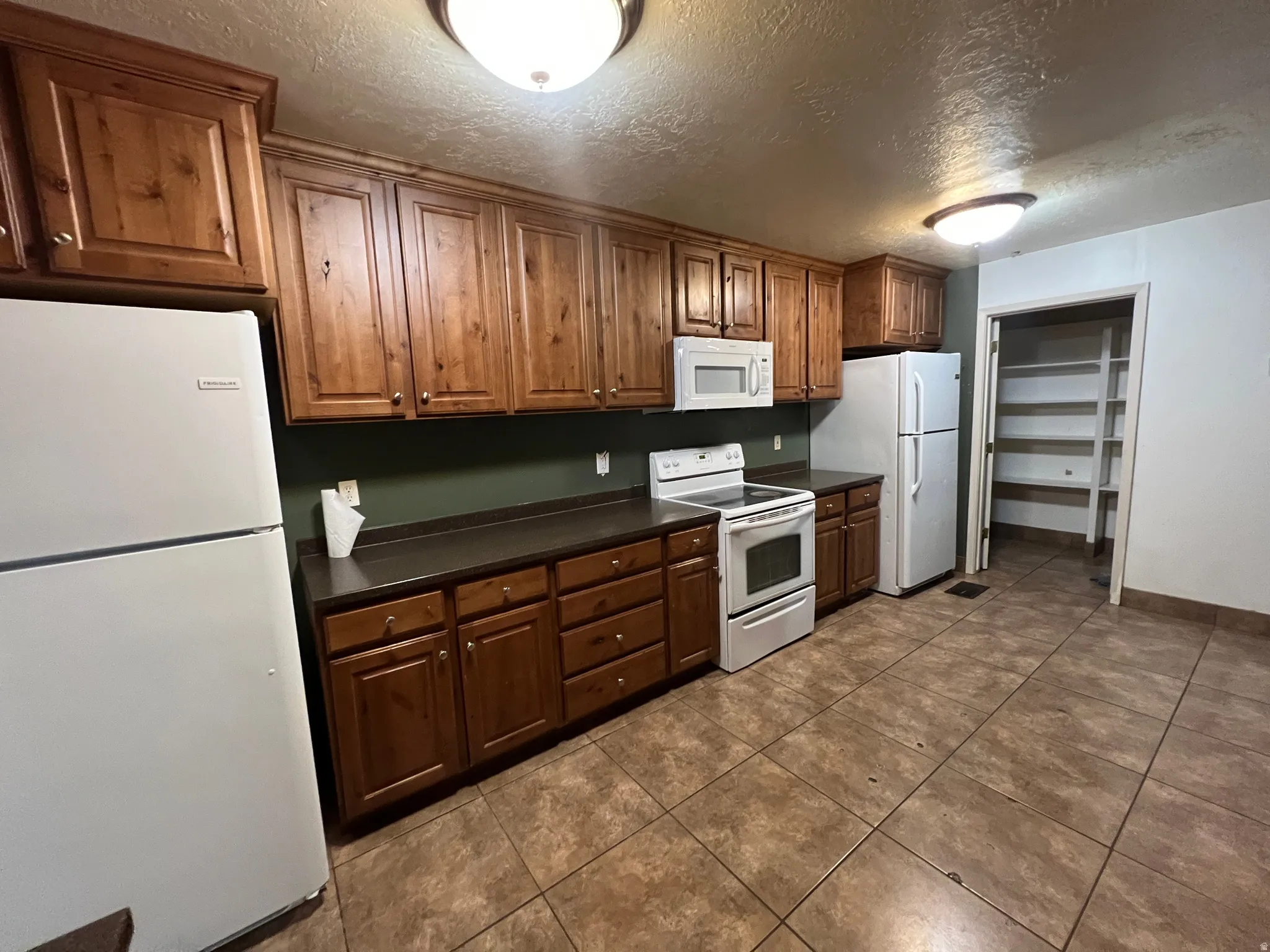 Kitchen featuring extensive wood cabinetry, dark countertops, white appliance suite, and tiled flooring