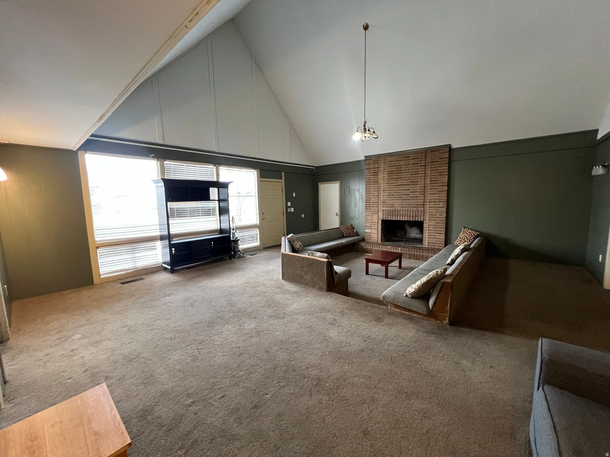 Spacious living area featuring a vaulted ceiling with exposed beams, an expansive brick fireplace, and large windows with horizontal blinds