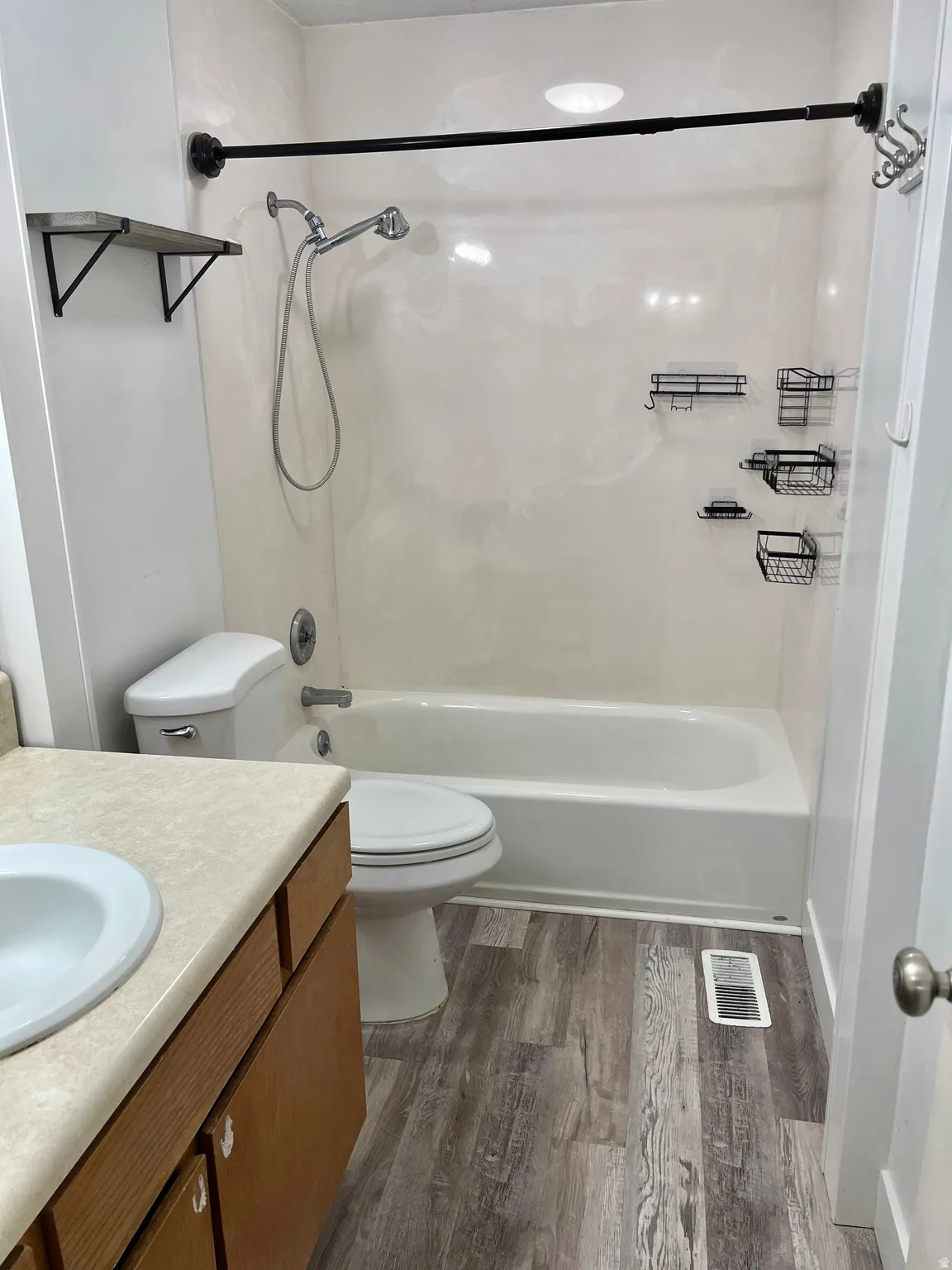 Full bathroom featuring a shower-over-tub combination with a wall-mounted showerhead, a toilet, a vanity with a drop-in sink and laminate countertop, and wood-finish flooring