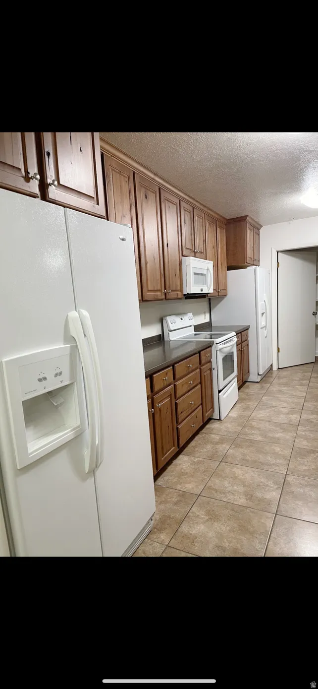 Kitchen featuring extensive wood cabinetry, white appliances, dark countertops, and tiled flooring
