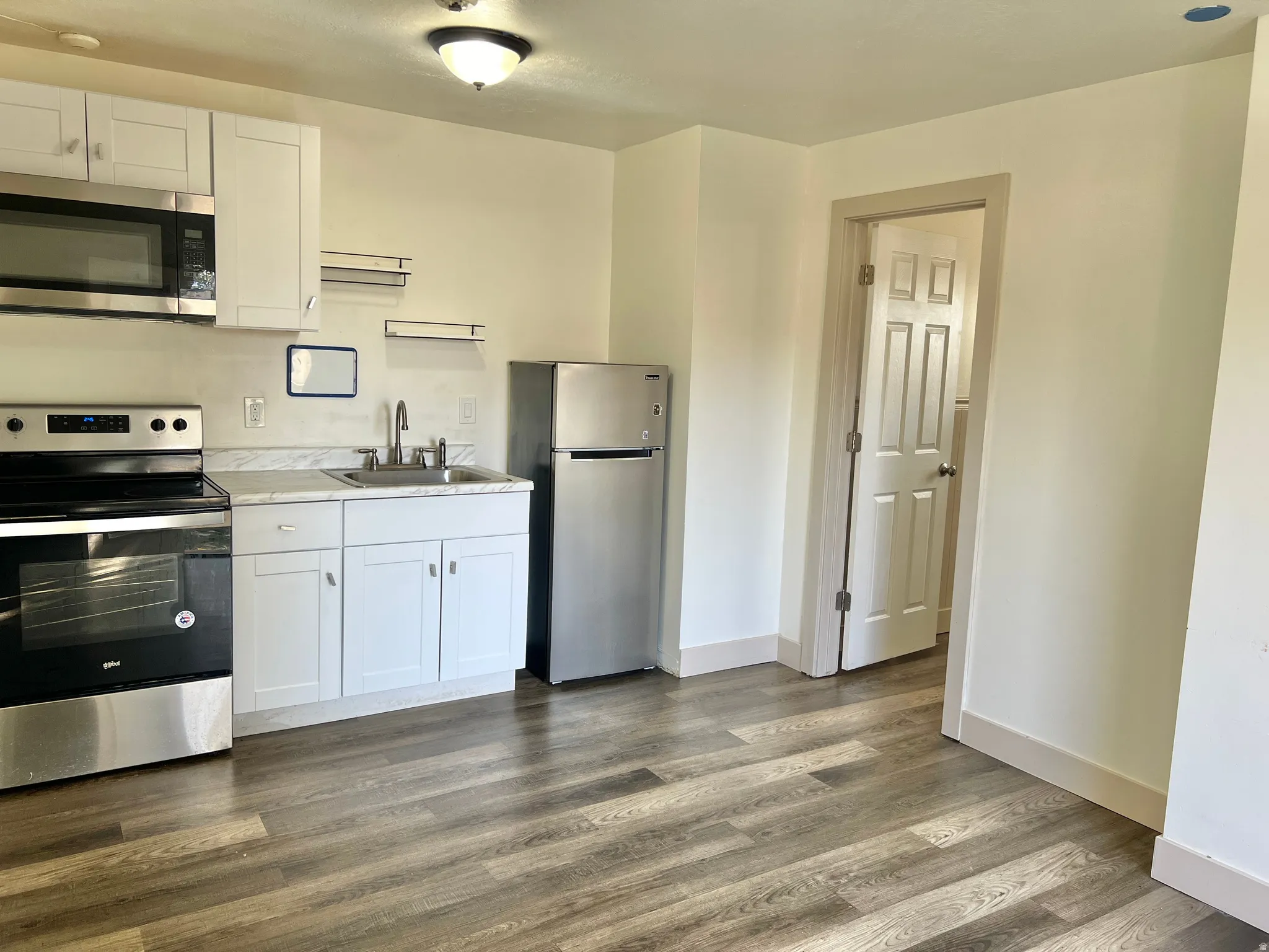 Kitchenette featuring a stainless steel appliance suite, white shaker cabinetry, light-toned countertops, wood-finish flooring, and a flush-mount ceiling light fixture