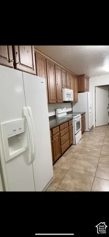 Kitchen featuring extensive wood cabinetry, white appliances, dark countertops, and tiled flooring