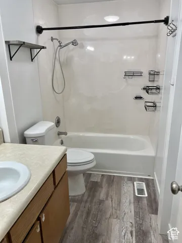 Full bathroom featuring a shower-over-tub combination with a wall-mounted showerhead, a toilet, a vanity with a drop-in sink and laminate countertop, and wood-finish flooring