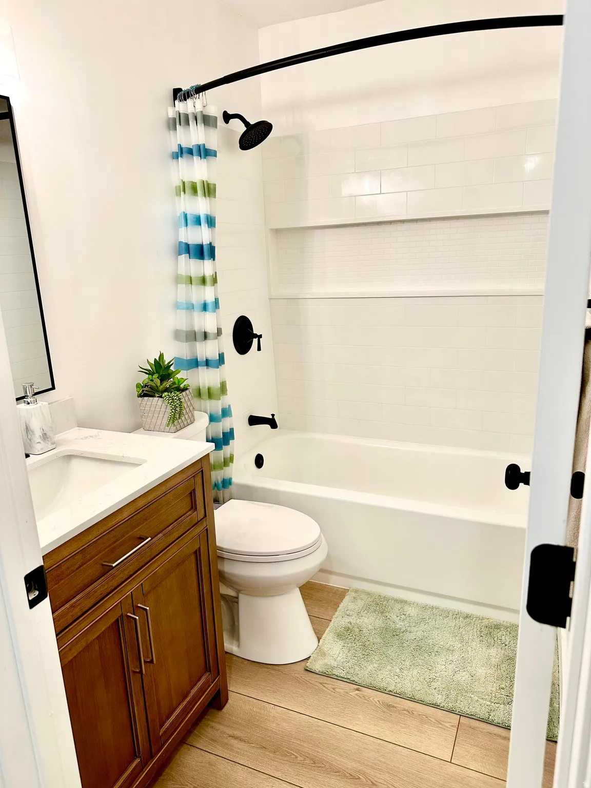 Bathroom featuring a white subway tile surround, matte black fixtures, a built-in shower shelf, a white vanity with a rectangular sink, and wood-finish flooring