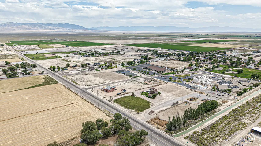 Expansive rural landscape featuring a paved road, agricultural fields, scattered residential structures, and distant mountains under a partially cloudy sky