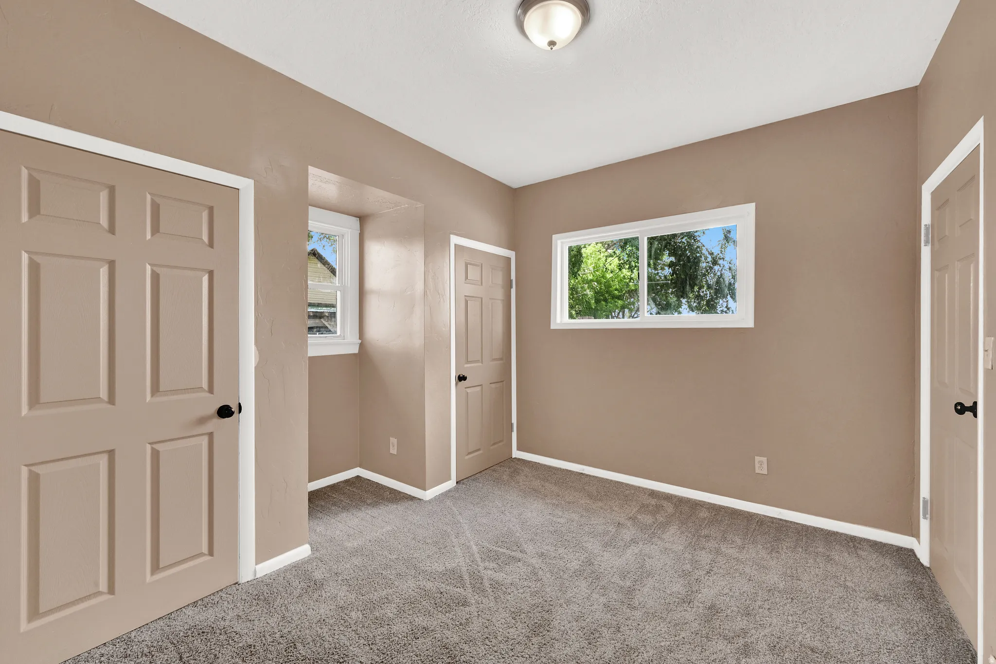 Neutral paint palette room featuring two windows with exterior foliage, gray carpet flooring, and white baseboards