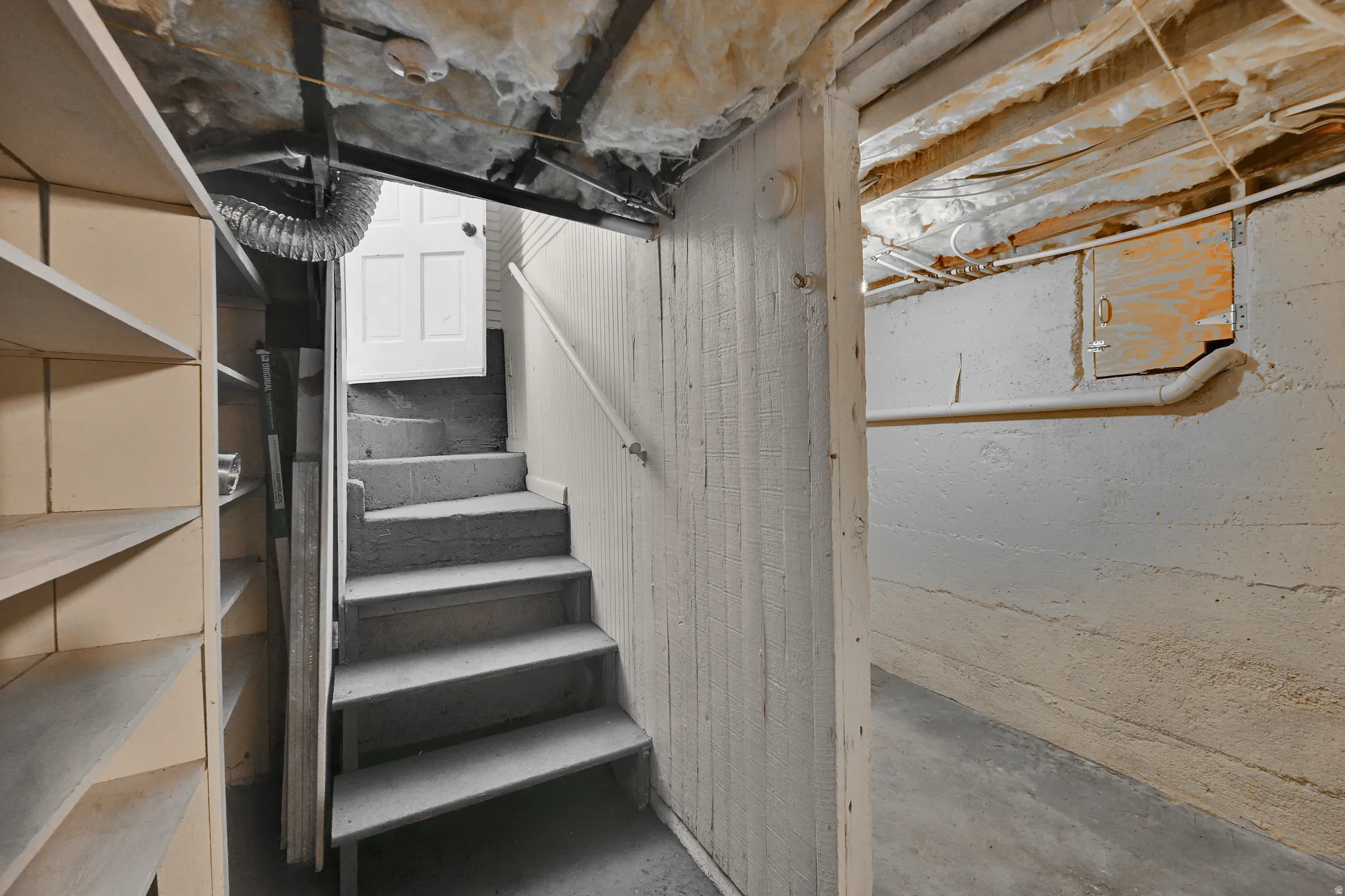 Concrete basement with exposed joists, insulated ceiling, built-in shelving, and a painted wood staircase with a handrail