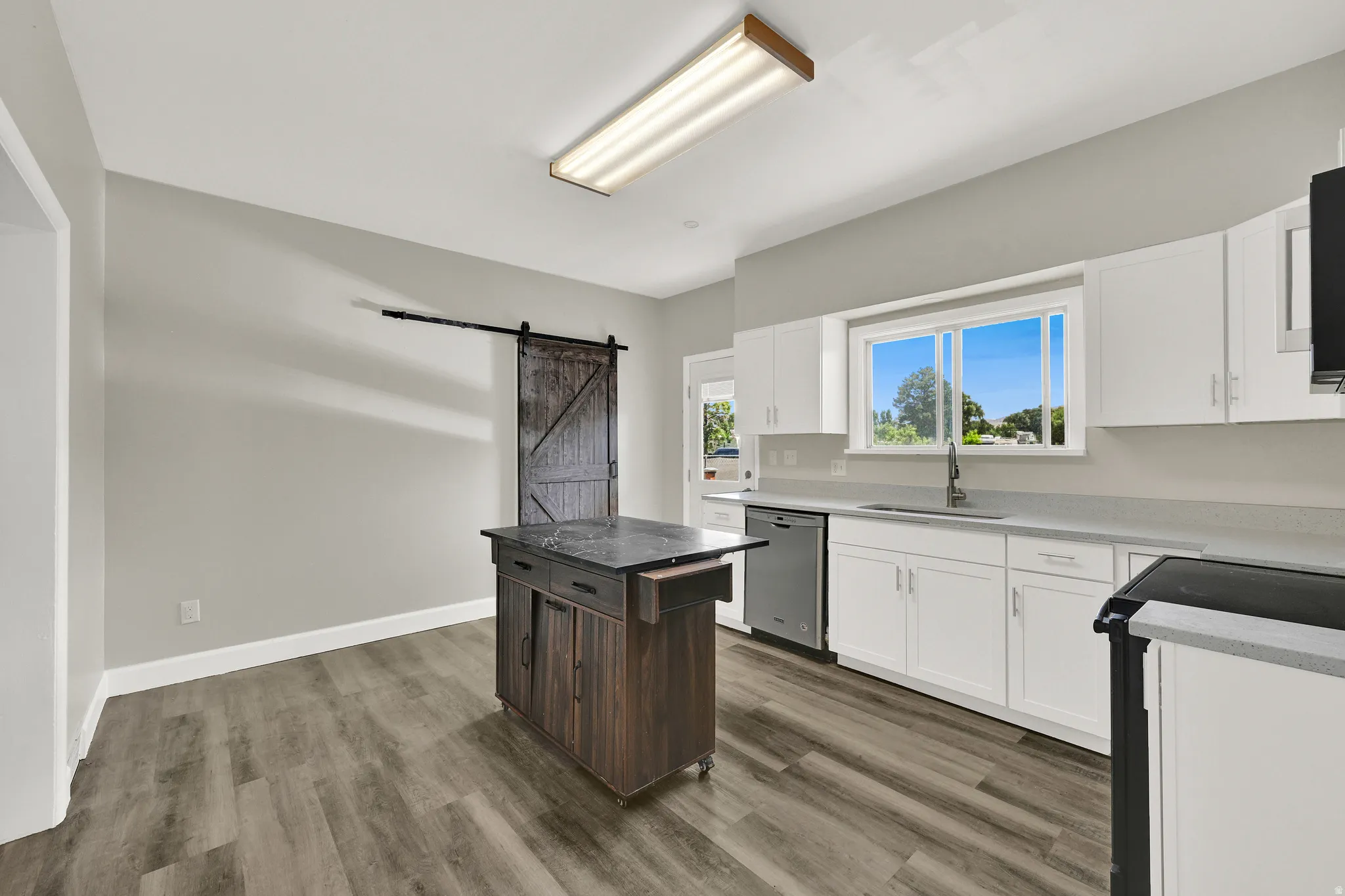 Kitchen featuring white cabinetry, light countertops, stainless steel dishwasher, wood-finish flooring, and a sliding barn door