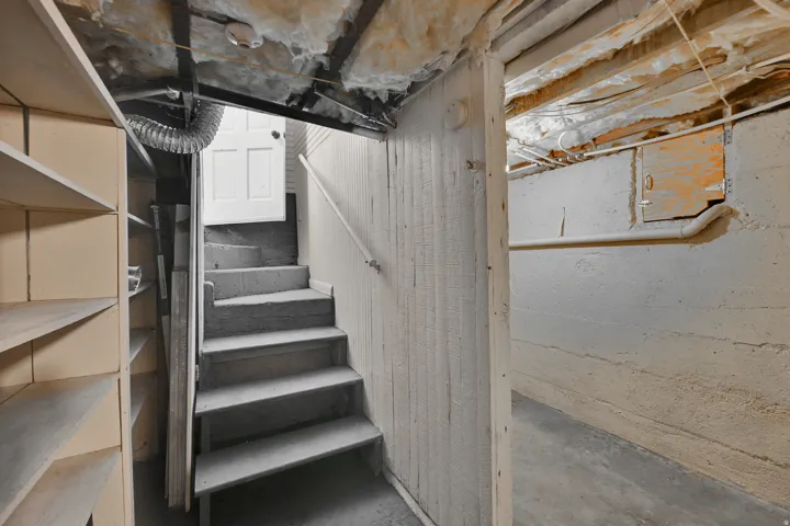 Concrete basement with exposed joists, insulated ceiling, built-in shelving, and a painted wood staircase with a handrail