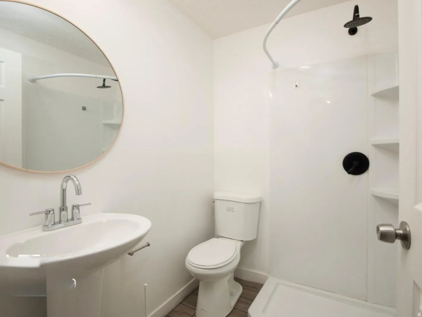 Pedestal sink with chrome faucet, circular mirror, toilet, and shower with black fixtures and integrated shelving