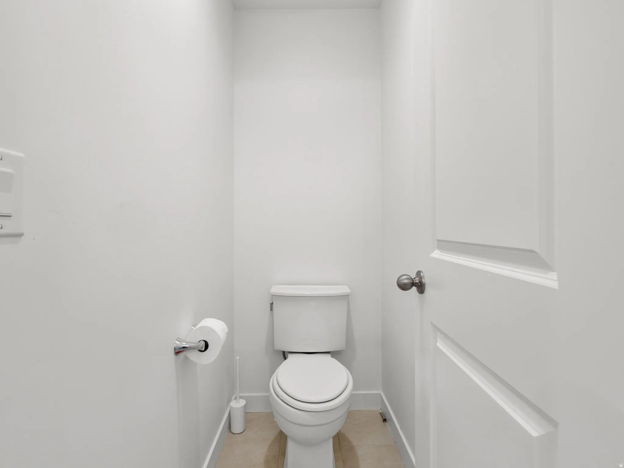 Brightly lit water closet featuring a contemporary toilet, light-colored tile flooring, and a panel-style door with a dark hardware finish