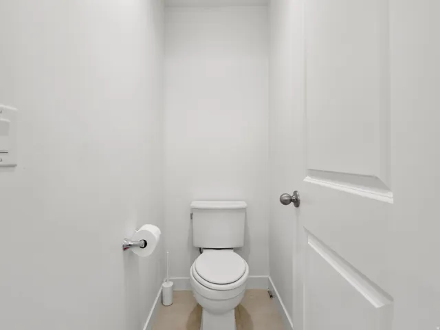 Brightly lit water closet featuring a contemporary toilet, light-colored tile flooring, and a panel-style door with a dark hardware finish