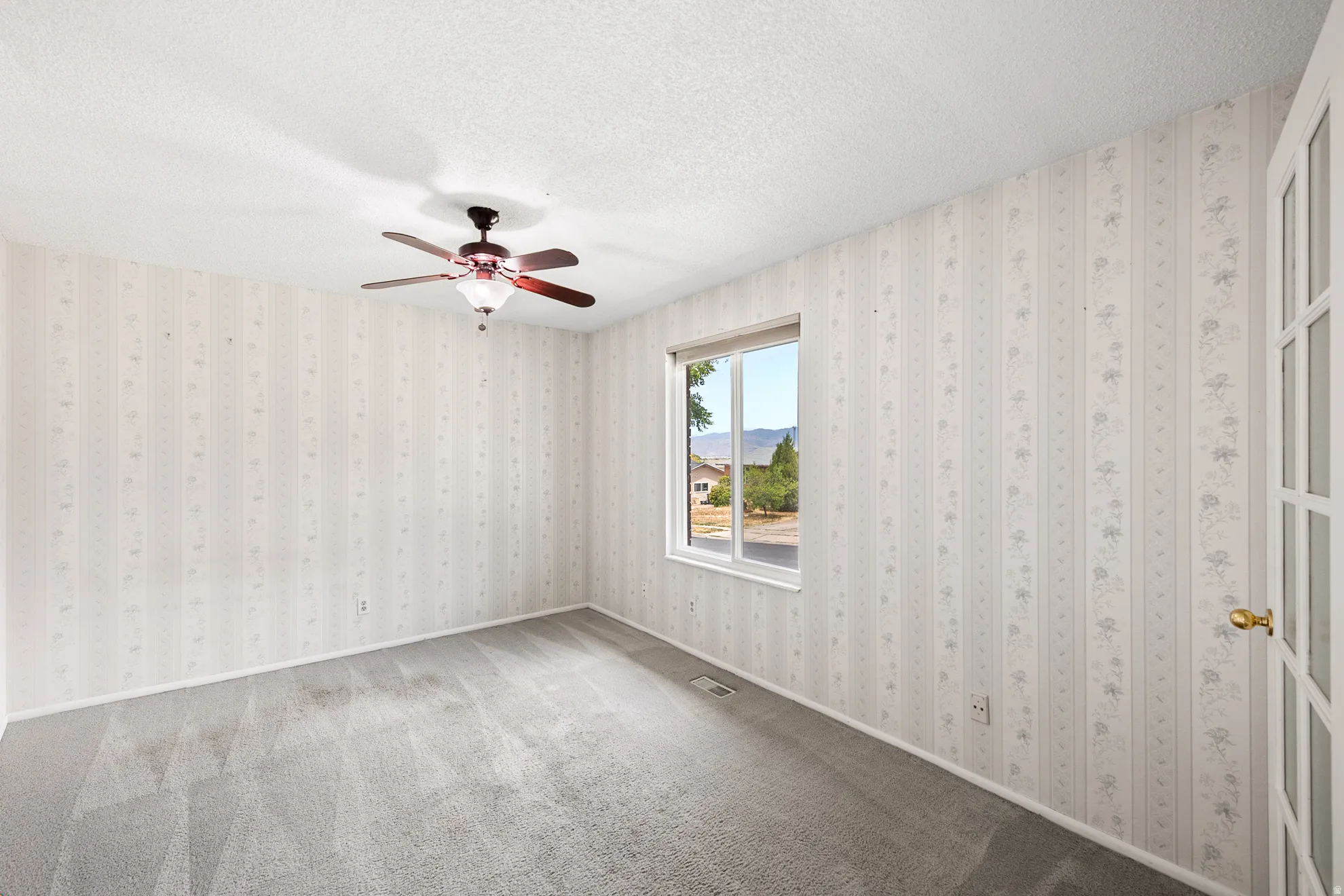 Spacious room featuring patterned wallpaper, neutral carpet, a ceiling fan with integrated lighting, and a window overlooking exterior landscaping