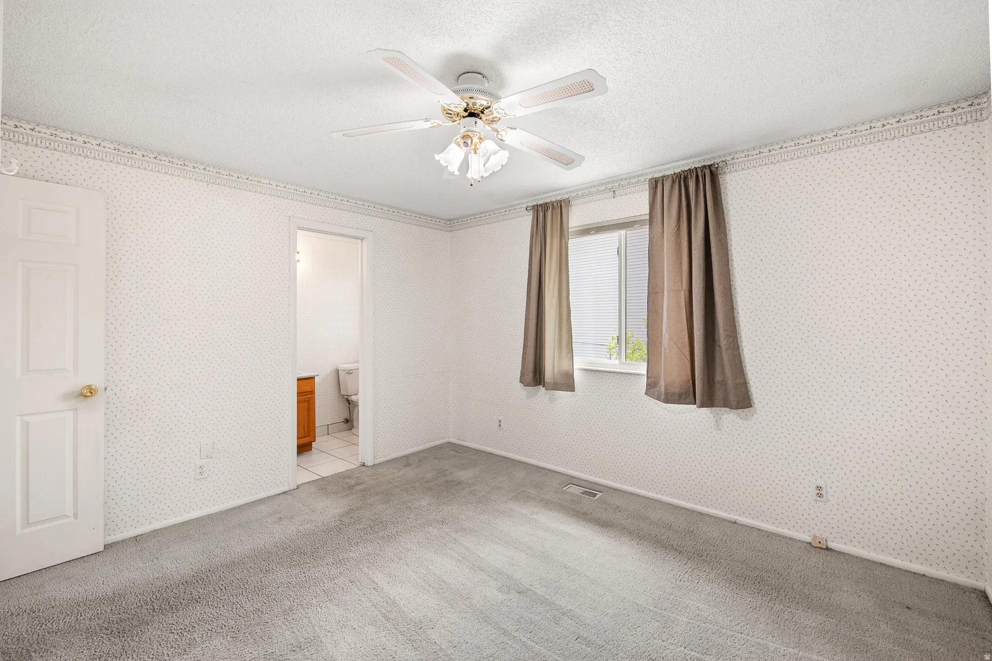 Carpeted room featuring a ceiling fan with light fixture, patterned wallpaper, a window with exterior visibility, and a doorway leading to a bathroom with a vanity
