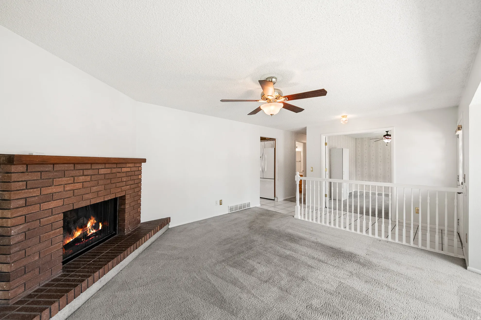 Spacious living area featuring a prominent brick fireplace with a wood mantel, light gray carpeting, and a ceiling fan with integrated lighting