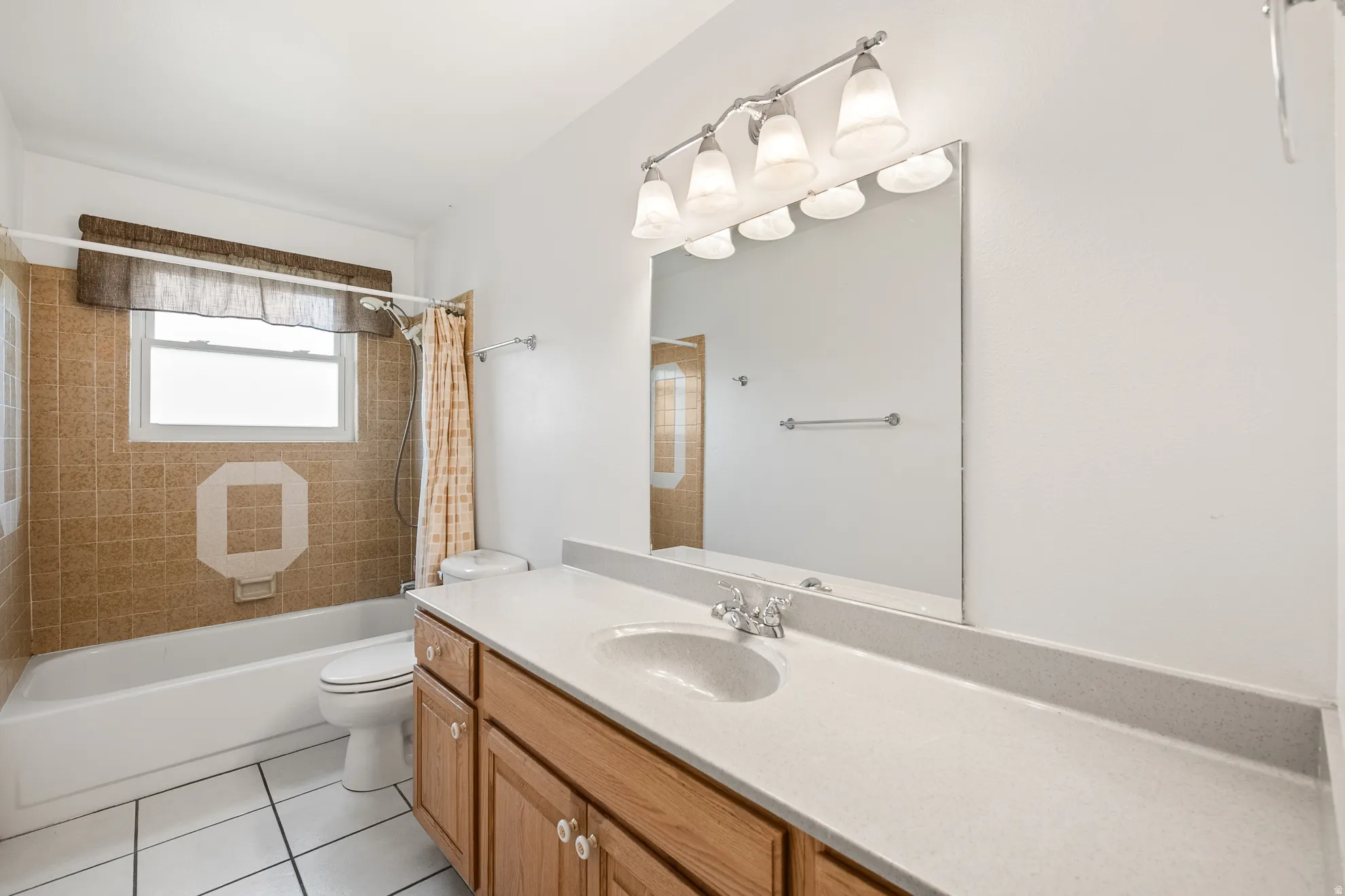 Bathroom featuring a white bathtub, a tiled surround with decorative accent, a window with a valance, a wood-finish vanity with a light-colored countertop, and a vanity mirror with an overhead light fixture